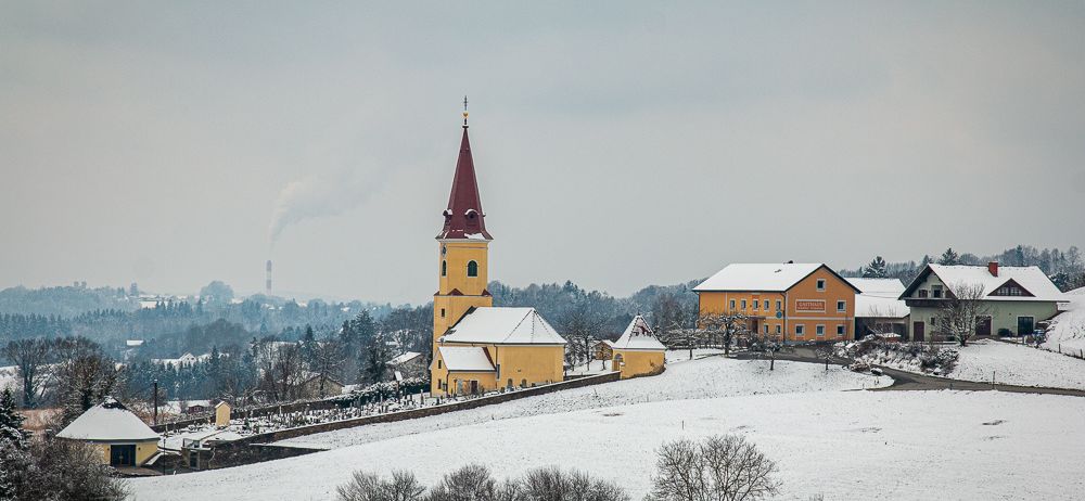 Eine Kirche mit einem Kirchturm steht in einer verschneiten Landschaft, umgeben von Häusern, Bäumen und einer Straße. Der Himmel ist bewölkt.