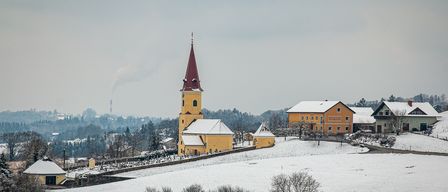 Eine Kirche mit einem Kirchturm steht in einer verschneiten Landschaft, umgeben von Häusern, Bäumen und einer Straße. Der Himmel ist bewölkt.
