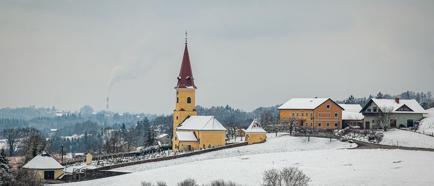 Eine Kirche mit einem Kirchturm steht in einer verschneiten Landschaft, umgeben von Häusern, Bäumen und einer Straße. Der Himmel ist bewölkt.