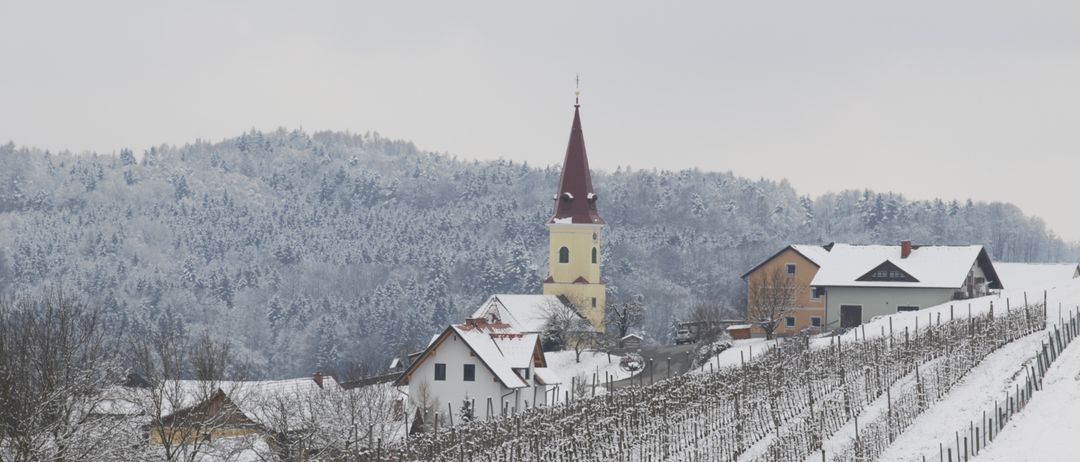 Eine verschneite Landschaft mit einem Kirchturm in der Mitte, umgeben von schneebedeckten Häusern und Bäumen. Die Häuser sind weiß und haben Schnee auf ihren Dächern.