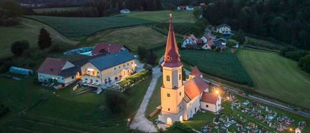 Ein Luftbild eines Dorfes bei Sonnenuntergang. Die Kirche hat einen Turm und ist von einem Friedhof umgeben. Es gibt Häuser und Bäume in der Ferne.