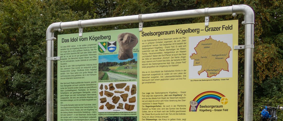 Ein Informationsschild am Koegelberg zeigt eine Idolstatue, historische Artefakte und eine Karte. Es betont die Bedeutung des Koegelbergs als kultureller und spiritueller Ort, mit Verweisen auf die katholische Kirche und biblische Geschichten.