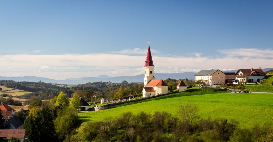 Eine Kirche mit einem roten Turm steht auf einem grünen Hügel, umgeben von einem Friedhof und Häusern. Der Himmel ist blau mit Wolken, und Berge sind in der Ferne.