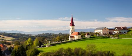 Eine Kirche mit einem roten Turm steht auf einem grünen Hügel, umgeben von einem Friedhof und Häusern. Der Himmel ist blau mit Wolken, und Berge sind in der Ferne.