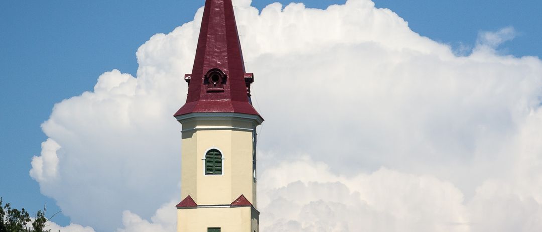 Ein hoher Kirchturm mit einem roten Dachfirst steht auffällig gegen einen blauen Himmel mit weißen Wolken, umgeben von Grün.