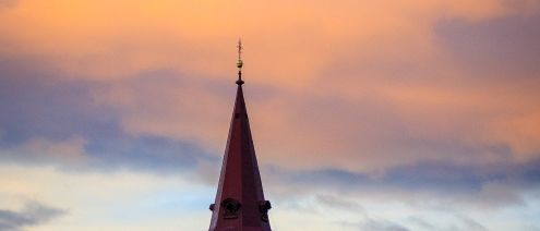 Eine Kirche mit einem hohen Turm steht in einer ländlichen Umgebung unter einem farbenfrohen Sonnenuntergangshimmel, umgeben von Bäumen und Gras.