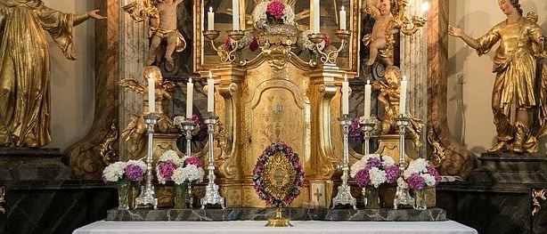 Ein kunstvoller Altar, geschmückt mit Kerzen, Blumen und Statuen. Zwei goldene Statuen flankieren den Altar auf beiden Seiten. Der Altar hat einen goldenen Rahmen und ein Bildnis eines Heiligen.