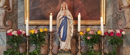 Ein Altar geschmückt mit einer Statue der Maria, Kerzen und Blumen, unter einem religiösen Gemälde und Cherubs. Der Altartisch hat ein weißes Tuch mit 'Heilige Maria, bitte für uns!' darauf geschrieben.