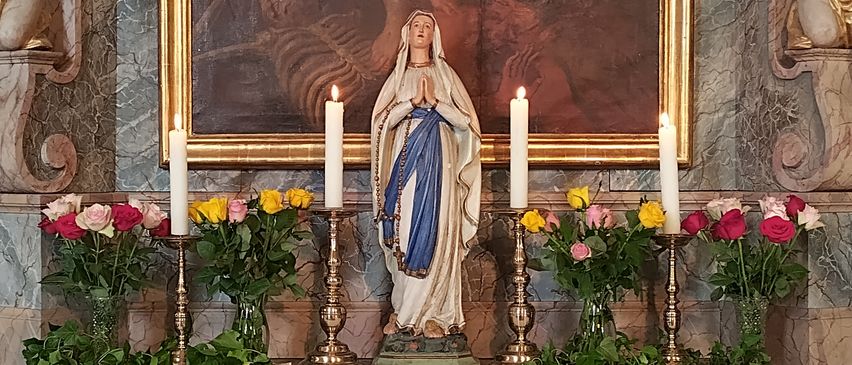 An altar adorned with a statue of Mary, candles, and flowers, beneath a religious painting and cherubs. The altar table has a white cloth with 'Holy Mary, please for us!' written on it.