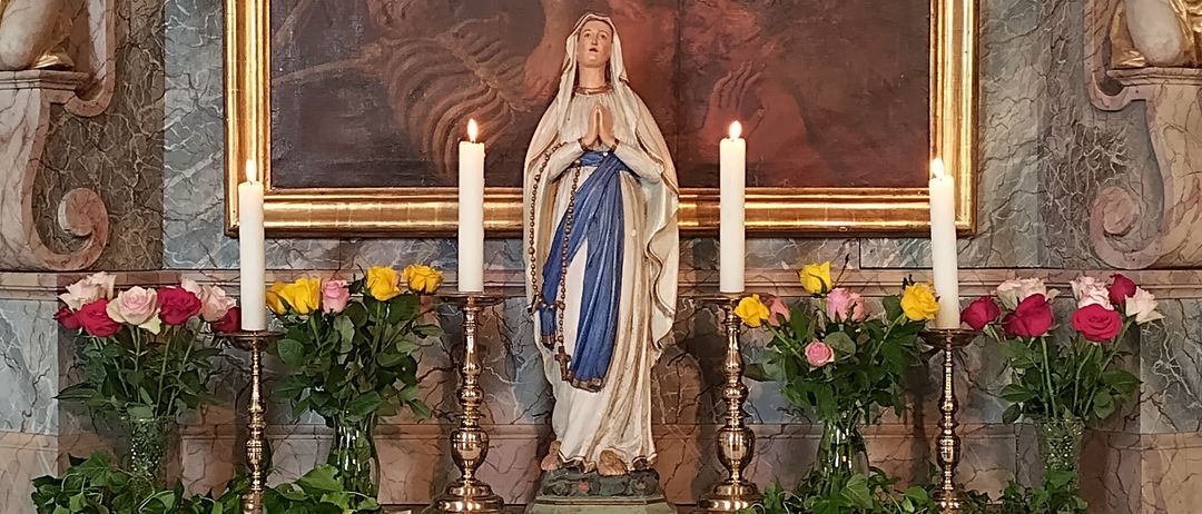 Ein Altar geschmückt mit einer Statue der Maria, Kerzen und Blumen, unter einem religiösen Gemälde und Cherubs. Der Altartisch hat ein weißes Tuch mit 'Heilige Maria, bitte für uns!' darauf geschrieben.