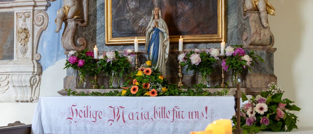 An altar adorned with flowers, candles, and a statue of the Virgin Mary. The altar cloth reads 'Heilige Maria, bitte für uns!'. Two cherub statues flank a large framed painting above the altar.