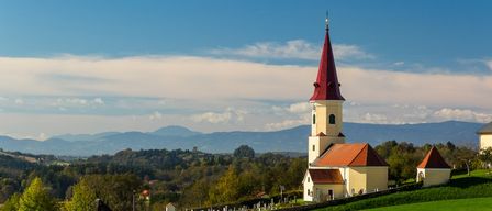 Eine malerische Kirche mit einem roten Turm steht auf einem Hügel, umgeben von einem grünen Friedhof und einem blauen Himmel mit Wolken.