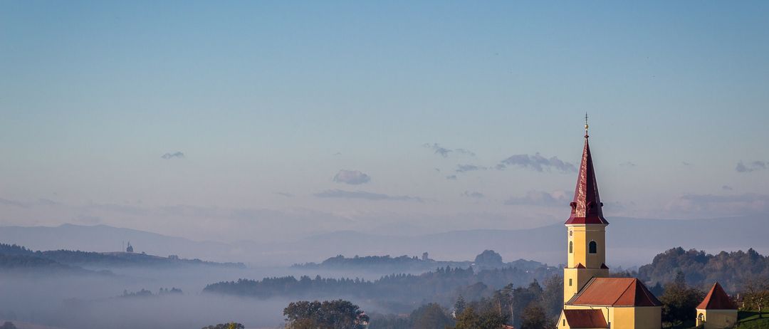 Eine gelbe Kirche mit einem roten Turm steht auf einem Hügel, umgeben von Nebel und Bäumen. Der Himmel ist blau mit einigen Wolken.