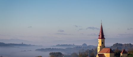 Eine gelbe Kirche mit einem roten Turm steht auf einem Hügel, umgeben von Nebel und Bäumen. Der Himmel ist blau mit einigen Wolken.