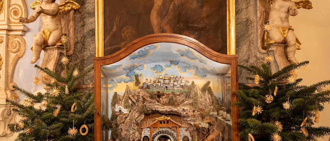 Eine detaillierte Krippenszene in einem Holzrahmen, ausgestellt in einer Kirche mit einem großen Gemälde im Hintergrund.