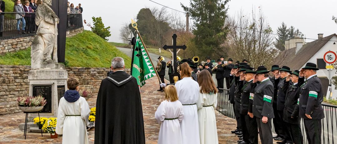 Eine feierliche Prozession mit einem Priester und Chor vor einem Kreuz, mit Zuschauern und einer Marschkapelle im Hintergrund.