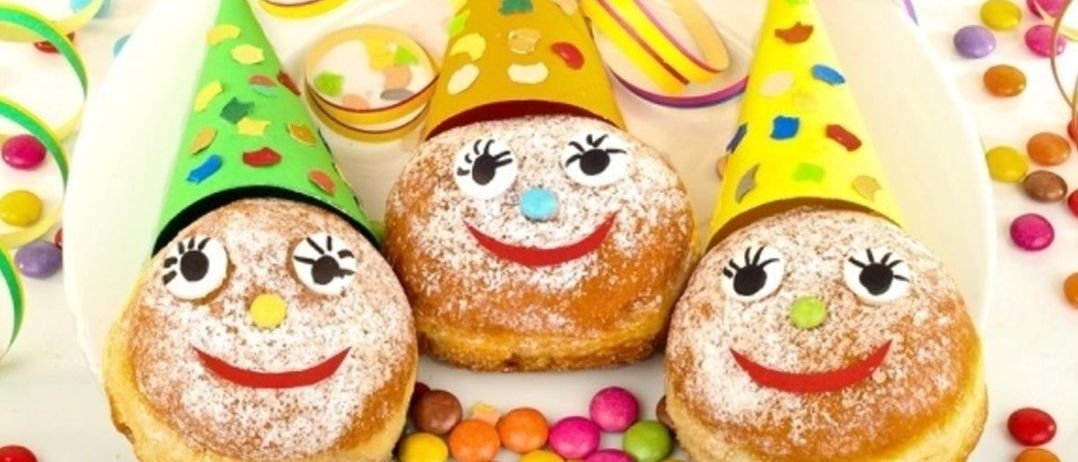 Drei Donuts mit bemalten Gesichtern und gelben Partyhüten stehen auf einem Teller mit bunten Süßigkeiten darum herum.