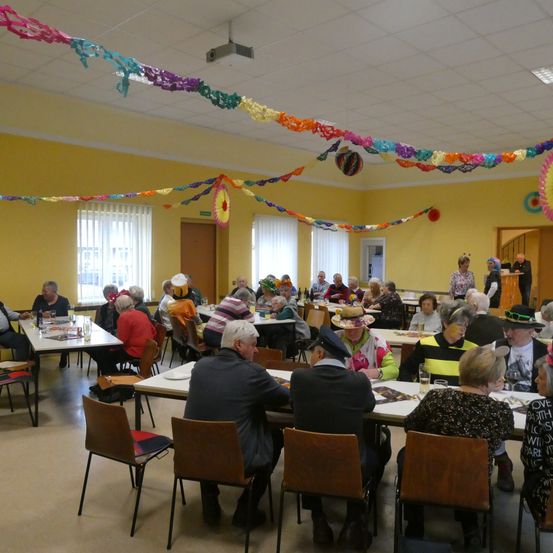 Ein großer Raum voller älterer Menschen, die an Tischen sitzen, einige tragen Hüte, und andere stehen in der Tür. Der Raum ist mit bunten Girlanden dekoriert, die von der Decke hängen.