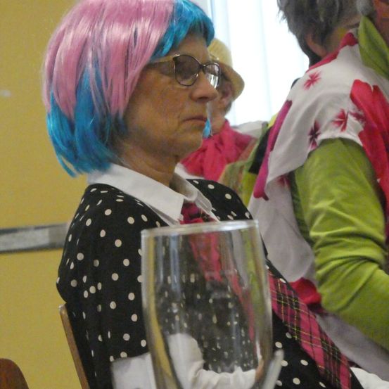 Eine ältere Frau mit rosa und blauen Haaren und einem gepunkteten Shirt sitzt an einem Tisch und hält ein Weinglas. Hinter ihr ist eine Person in einem grünen und weißen Blumenhemd zu sehen.