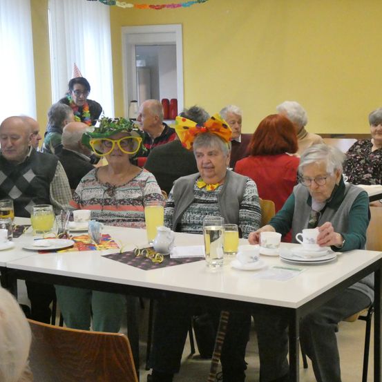 Eine Gruppe älterer Menschen sitzt um einen Tisch in einem Raum. Einige tragen Hüte und Brillen. Auf dem Tisch stehen Teller, Tassen und Gläser. Der Raum hat gelbe Wände und weiße Vorhänge.