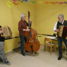 Drei Männer spielen Instrumente in einem Raum. Der Mann auf der linken Seite spielt Akkordeon. Der Mann in der Mitte spielt Cello. Der Mann auf der rechten Seite spielt Akkordeon.