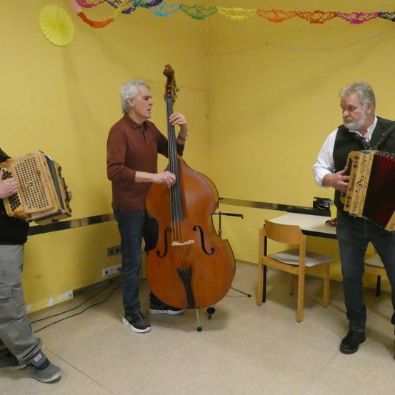 Drei Männer spielen Instrumente in einem Raum. Der Mann auf der linken Seite spielt Akkordeon. Der Mann in der Mitte spielt Cello. Der Mann auf der rechten Seite spielt Akkordeon.