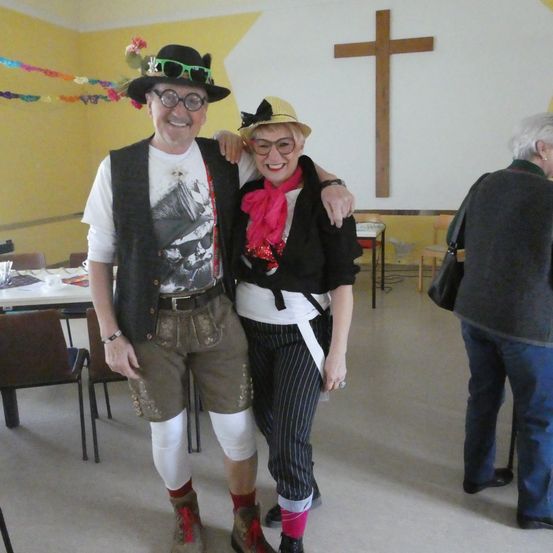 Zwei Personen in traditioneller bayerischer Kleidung stehen lächelnd in einem Raum mit einem Holzkreuz an der Wand. Hinter ihnen steht ein Tisch mit Stühlen, und eine weitere Person ist teilweise sichtbar.