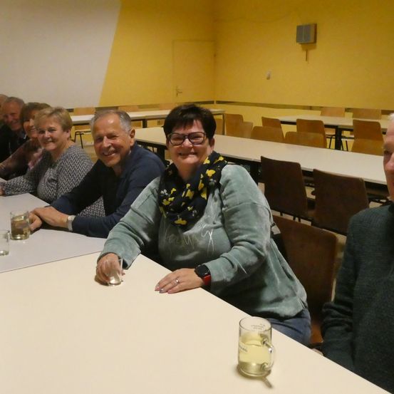 Gruppe lächelnder Menschen, die mit Gläsern an einem Tisch in einem gelb gestrichenen Raum mit vielen leeren Stühlen sitzen.