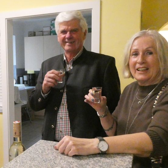 Ein älterer Mann und eine ältere Frau stehen in einer Küche, lächeln und halten kleine Gläser. Eine Flasche ist auf dem Tresen.