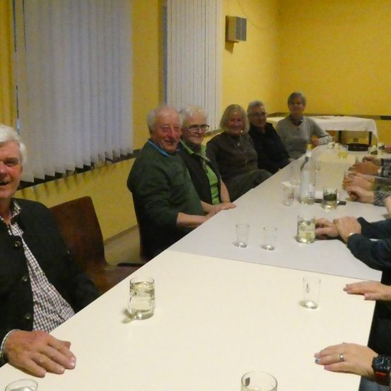 Eine Gruppe älterer Erwachsener sitzt um einen Tisch mit Gläsern und Flaschen. Sie lächeln und scheinen ein Gespräch zu genießen. Der Raum hat gelbe Wände und weiße Jalousien.