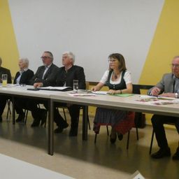 Sieben Menschen sitzen an einem Tisch in einem Raum. Vier Männer und drei Frauen. Eine Frau trägt ein Dirndl. Alle sehen ernst aus.