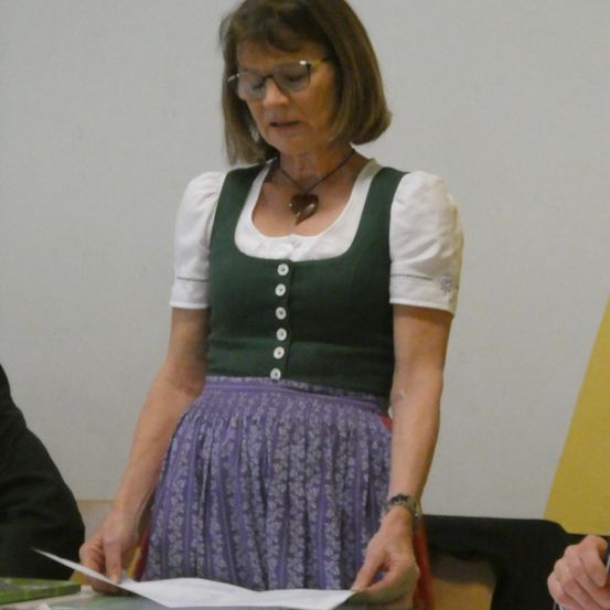 Eine Frau in traditioneller Kleidung steht an einem Tisch, hält ein Papier und scheint zu lesen. Sie trägt eine Brille, eine Halskette und ein weißes Hemd mit grüner Weste.