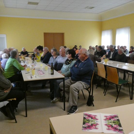 Eine Gruppe älterer Menschen sitzt um Tische in einem Raum. Die Tische haben Gläser, Flaschen und andere Gegenstände. Der Raum hat gelbe Wände und weiße Decken.