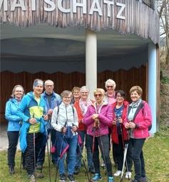 Eine Gruppe von Menschen in Winterkleidung steht auf einer Rasenfläche mit Wanderstöcken. Sie posieren für ein Foto vor einem Gebäude mit einem Holzzaun. Das Schild lautet 'MATSCHATZ'.
