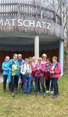 Eine Gruppe von Menschen in Winterkleidung steht auf einer Rasenfläche mit Wanderstöcken. Sie posieren für ein Foto vor einem Gebäude mit einem Holzzaun. Das Schild lautet 'MATSCHATZ'.
