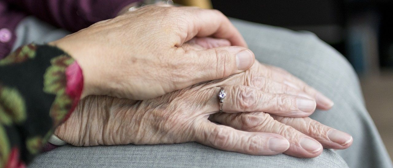 Die faltigen Hände einer älteren Frau sind ineinander verschränkt, mit einem Ring am Finger, auf einer grauen Oberfläche.