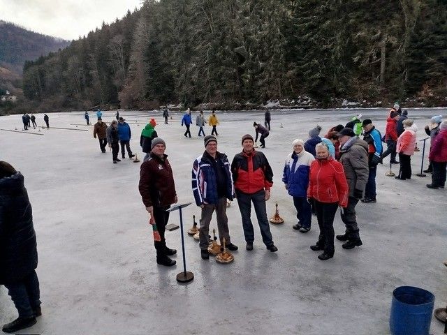Eine Gruppe von Menschen steht auf einem zugefrorenen See, möglicherweise spielen sie ein Spiel. Einige von ihnen tragen Mützen und Schuhe. Es gibt Objekte, die wie Glocken aussehen, auf dem Boden.