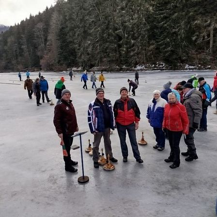 Eine Gruppe von Menschen steht auf einem zugefrorenen See, möglicherweise spielen sie ein Spiel. Einige von ihnen tragen Mützen und Schuhe. Es gibt Objekte, die wie Glocken aussehen, auf dem Boden.