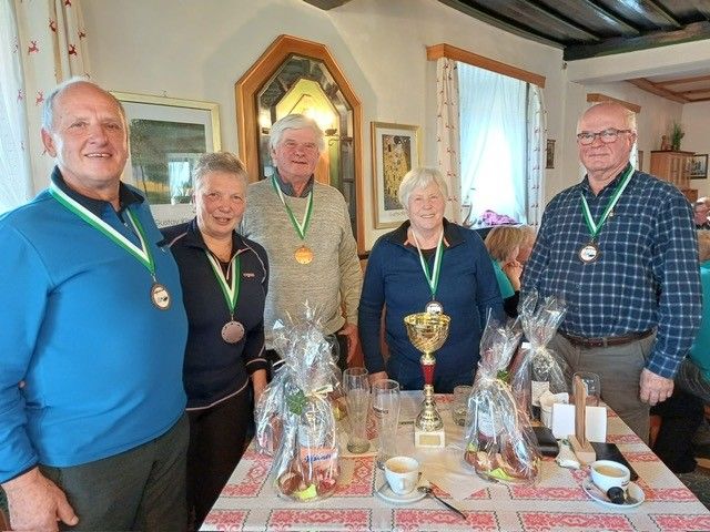 Fünf Personen mit Medaillen posieren für ein Foto bei einer Zusammenkunft, stehend hinter einem Tisch mit Trophäen und verpackten Geschenken. Zwei Tassen mit Untertassen stehen auf dem Tisch, mit zusätzlichen Personen im Hintergrund.
