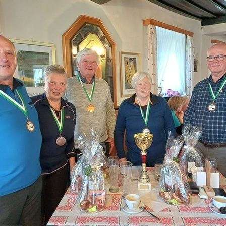 Fünf Personen mit Medaillen posieren für ein Foto bei einer Zusammenkunft, stehend hinter einem Tisch mit Trophäen und verpackten Geschenken. Zwei Tassen mit Untertassen stehen auf dem Tisch, mit zusätzlichen Personen im Hintergrund.