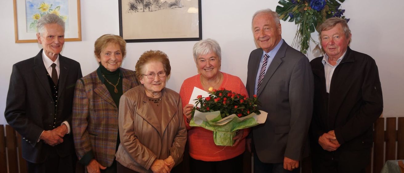 Eine Gruppe von Menschen steht drinnen zusammen. Eine ältere Frau hält einen Blumenstrauß. Die anderen lächeln. Ein gerahmtes Bild und eine Blumendekoration hängen an der Wand.