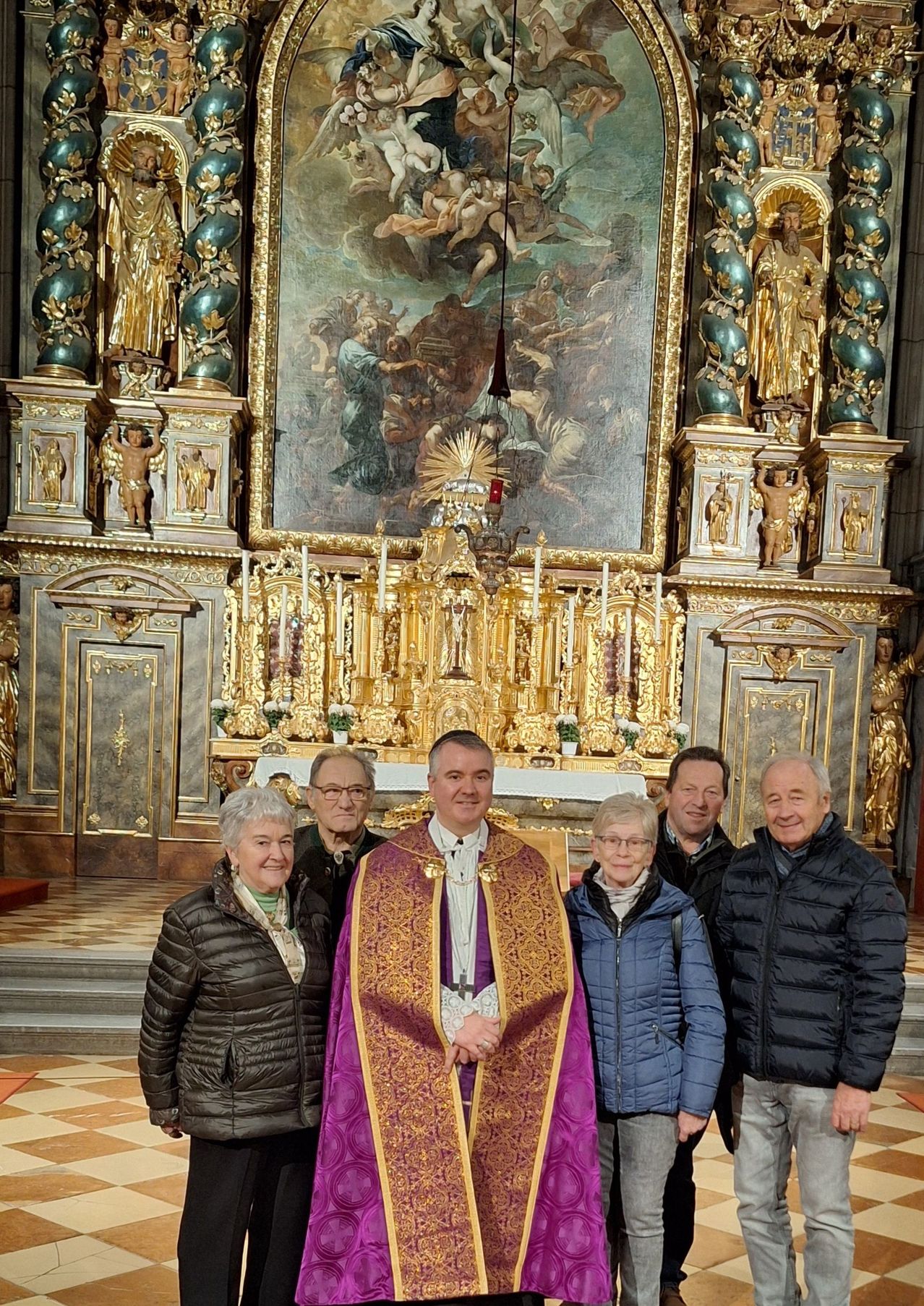Eine Gruppe von Menschen steht vor einem Kirchenaltar, ein Priester in zeremoniellen Gewändern. Dahinter ist ein großes Gemälde zu sehen, und Statuen befinden sich auf beiden Seiten des Altars.
