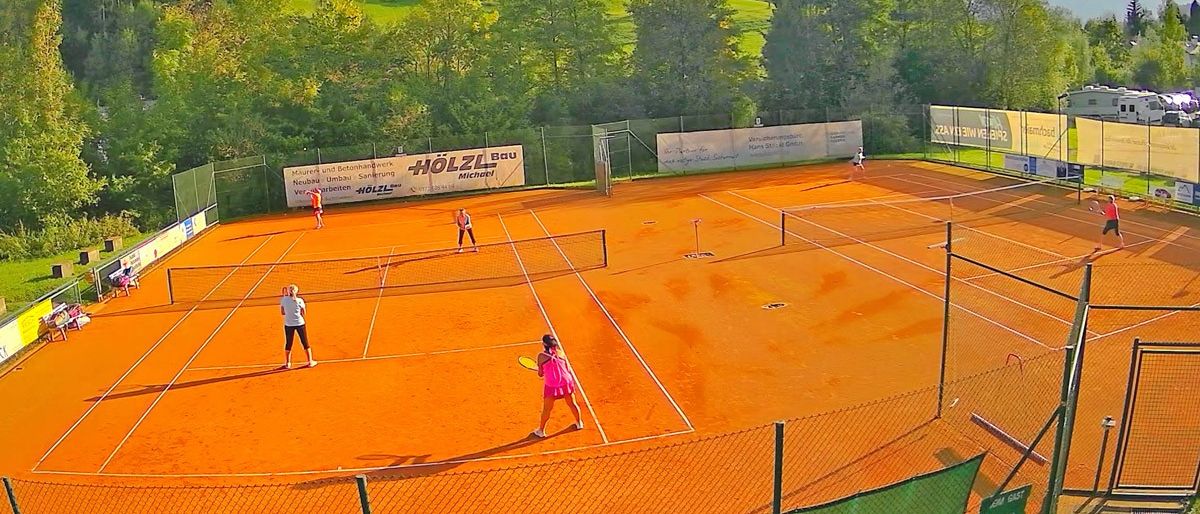 Eine Luftaufnahme eines Tennisplatzes mit vier Spielern. Drei spielen ein Match, einer geht zum Netz. Banner sind an der Umzäunung sichtbar.