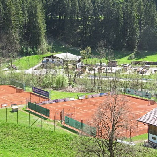 Luftaufnahme eines Tennisplatzes in einem grünen Tal, umgeben von Bäumen und einem Haus.