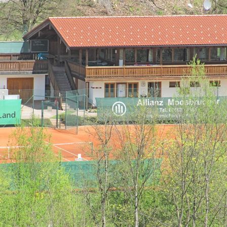 Ein großes Haus mit rotem Dach und mehreren Fenstern befindet sich in einem grünen Bereich, mit einem Tennisplatz und einem Schild davor.