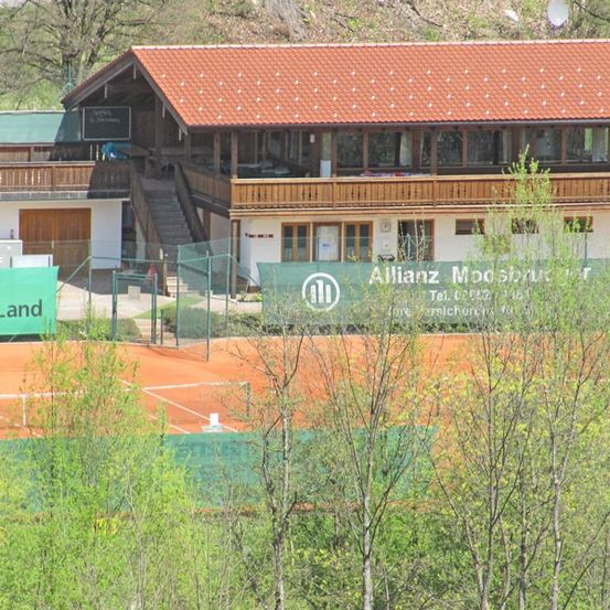 Ein großes Haus mit rotem Dach und mehreren Fenstern befindet sich in einem grünen Bereich, mit einem Tennisplatz und einem Schild davor.