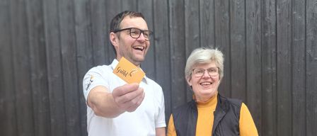 Ein Mann und eine Frau stehen vor einer Holzwand und lächeln. Der Mann hält eine gelbe Karte und die Frau trägt eine Brille.