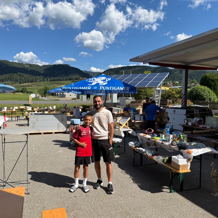 Zwei Personen stehen bei einem Tisch bei einer Outdoor-Veranstaltung. Ein Mann und ein Junge halten Händchen. Hinter ihnen sind Tische mit verschiedenen Gegenständen aufgestellt, und ein blauer Zelt mit Solarpanelen ist zu sehen. In der Ferne sind Berge und ein klarer Himmel zu sehen.