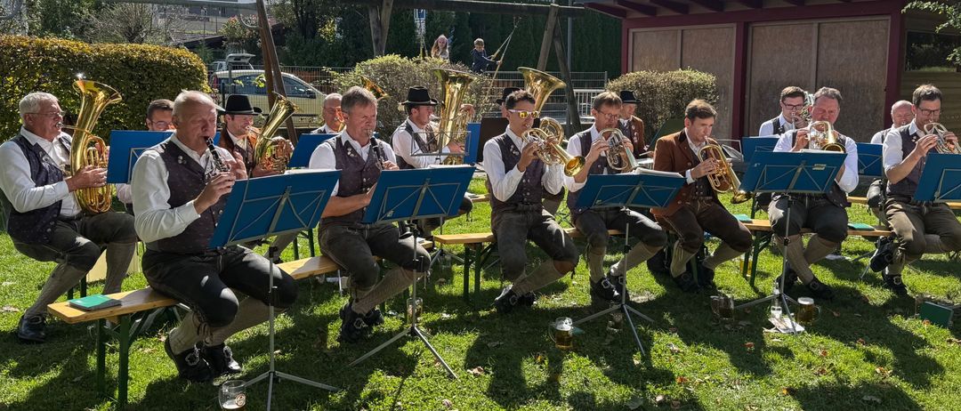 Eine Brassband in traditioneller Kleidung spielt auf einem Rasen mit Bergen im Hintergrund. Bänke, Notenständer und ein Gebäude sind sichtbar.