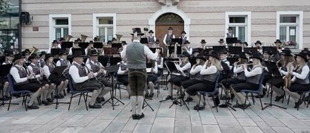 Ein Musikensemble in traditioneller Kleidung tritt auf einer Bühne vor einem Gebäude auf, mit Datum und Ort für bevorstehende Konzerte.